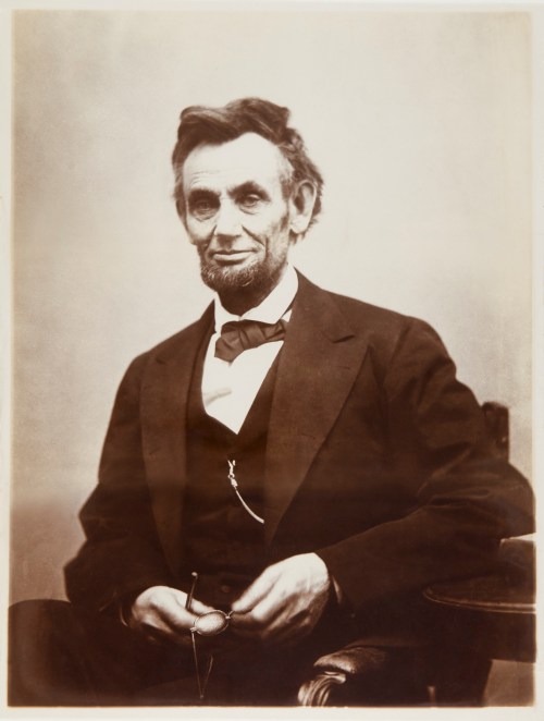 A historical portrait of a man with a beard, wearing a suit and bow tie, seated with glasses in hand.