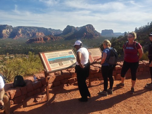 Hikers reading informational signs with a scenic view of red rock formations and mountains in the background.