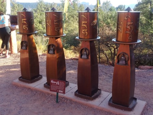 Four prayer wheels on wooden stands, each adorned with symbols, set in a natural outdoor environment.
