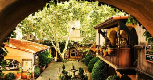 A charming courtyard with lush greenery, potted plants, and rustic architecture under a sunny sky.