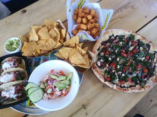 A spread of food including tacos, chips, guacamole, a salad, tater tots, and a colorful pizza on a wooden table.