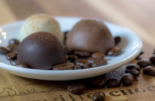 A white plate with three chocolate truffles surrounded by coffee beans on a wooden surface.
