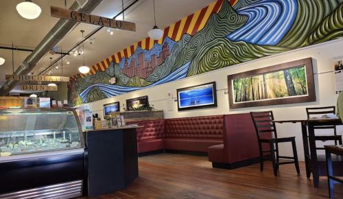 A cozy café interior featuring a colorful mural, seating area, and display case with gelato.