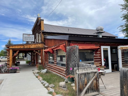 A rustic wooden building with a sign reading "Bison's Bounty," featuring a porch and flower pots outside.