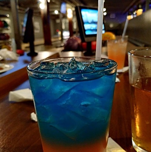 A colorful layered drink with blue and orange hues, served in a glass with ice and a straw, in a bowling alley setting.