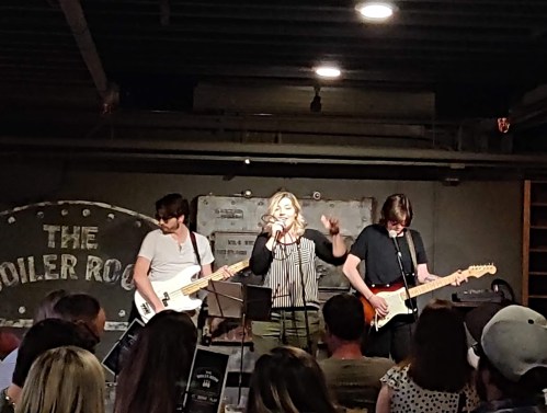 A live band performs on stage at a venue, with a singer and two guitarists in front of an audience.