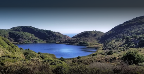 A serene landscape featuring a blue lake surrounded by green hills and distant ocean views under a clear sky.
