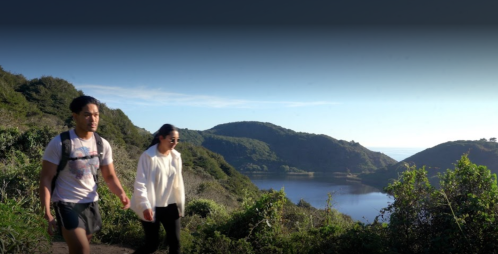 Two hikers walk along a scenic trail with a lake and hills in the background under a clear blue sky.