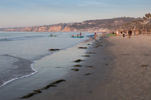 A serene beach scene with people walking, kayaking, and gentle waves lapping at the shore under a soft sky.