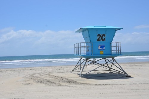 A blue lifeguard tower labeled "2C" on a sandy beach with calm waves and a clear sky in the background.