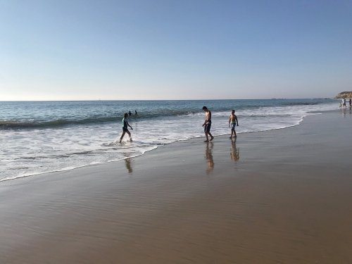 People walking along a sandy beach, with gentle waves lapping at the shore under a clear blue sky.