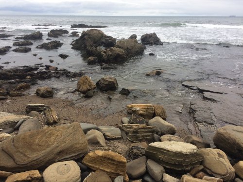 Rocky shoreline with waves gently crashing against the stones under a cloudy sky.