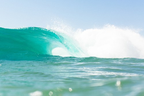 A close-up of a turquoise ocean wave curling, with white foam and a clear blue sky in the background.
