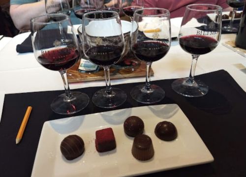 Four glasses of red wine lined up beside a plate of assorted chocolates, including truffles and a square chocolate.