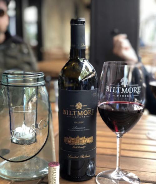 A bottle of Biltmore Malbec wine next to a glass of red wine on a wooden table, with a cork and jar in the background.
