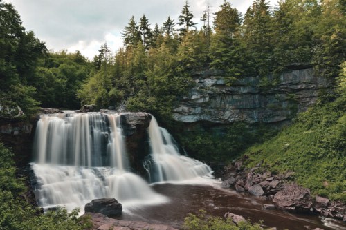 A serene waterfall cascades over rocky cliffs, surrounded by lush green trees and a cloudy sky.