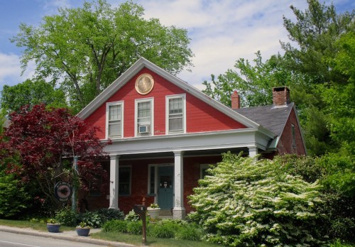 A charming red house with a porch, surrounded by lush greenery and colorful plants under a blue sky.