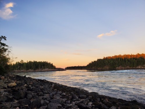 A serene river scene at sunset, with rocky shores and trees reflecting warm colors in the water.