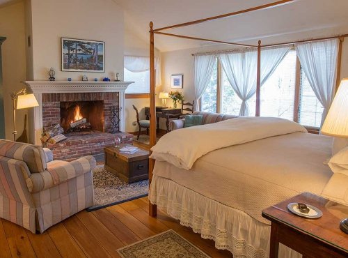 Cozy bedroom with a four-poster bed, fireplace, and comfortable seating by large windows. Warm, inviting atmosphere.