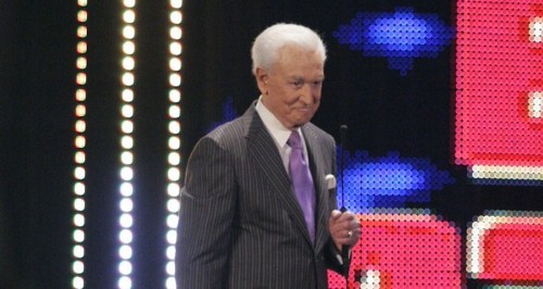 A man in a pinstripe suit and purple tie stands on stage, holding a microphone, with colorful lights in the background.