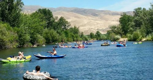 A busy river scene with people floating on colorful tubes and kayaks, surrounded by trees and mountains in the background.