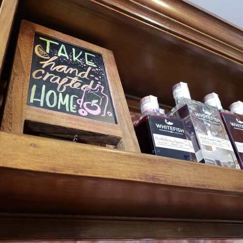 A wooden sign reads "Take handcrafted home" above bottles labeled "Whitefish" on a shelf.