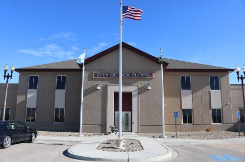 City of Box Elder building with an American flag in front, clear blue sky, and accessible parking spaces.