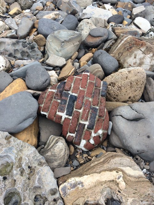 A heart-shaped piece of brick among various smooth stones on a rocky beach.