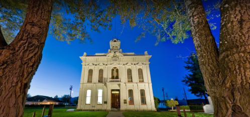 Historic building illuminated at dusk, framed by trees, with a clear blue sky in the background.