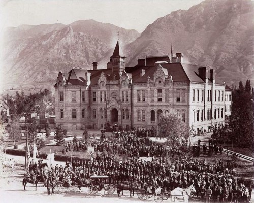 Historic black-and-white photo of a large crowd in front of a grand building, with mountains in the background.