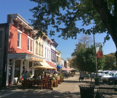 Charming street scene with colorful storefronts, outdoor seating, and trees under a clear blue sky.