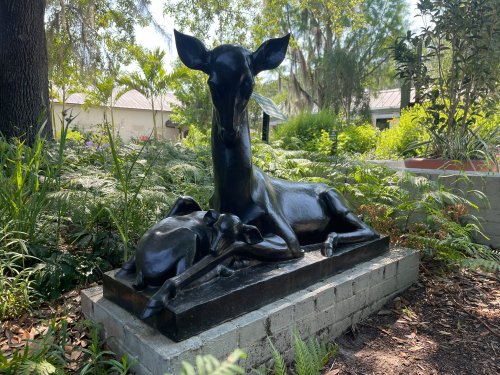 A black sculpture of a deer lying beside a dog, surrounded by lush greenery and plants.