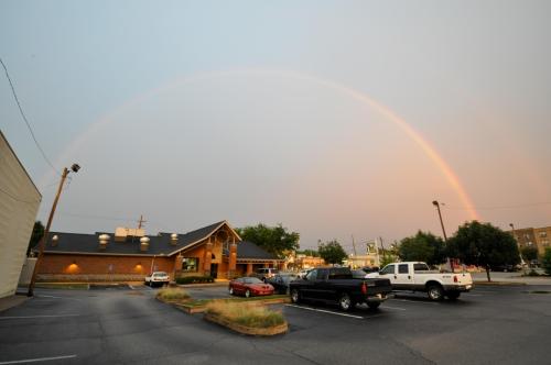 A rainbow arcs over a parking lot with cars, near a restaurant and shops under a cloudy sky.