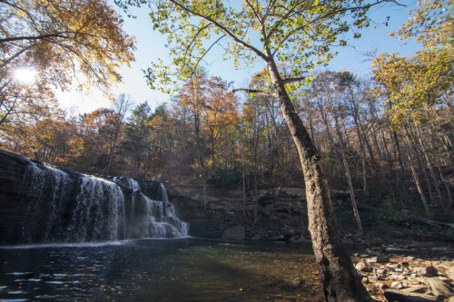 A serene waterfall cascades into a calm pool, surrounded by autumn foliage and trees under a clear blue sky.