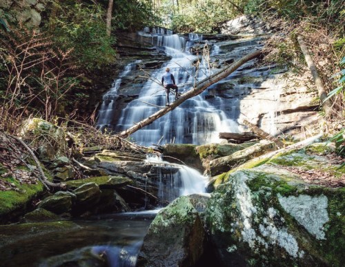A person stands on rocks near a waterfall, surrounded by lush greenery and flowing water.