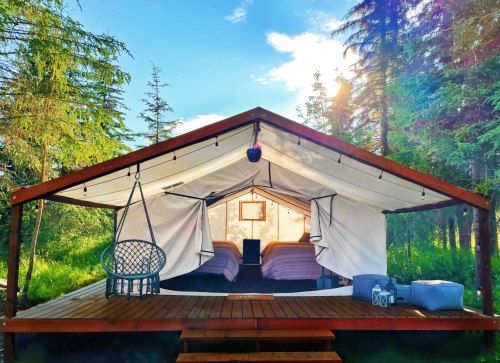 A cozy glamping tent with two beds, surrounded by trees and sunlight, featuring a wooden deck and outdoor seating.