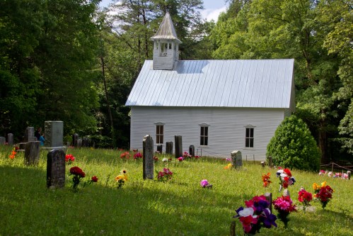 A white church with a steeple surrounded by a cemetery adorned with colorful flowers in a lush green setting.