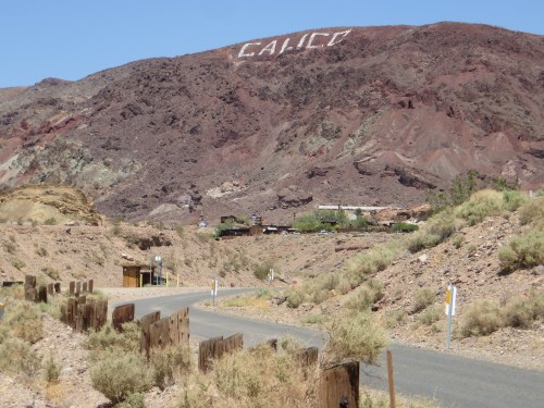 A desert landscape with a road leading to a mountain that has "CALICO" written on it, surrounded by rocky terrain.