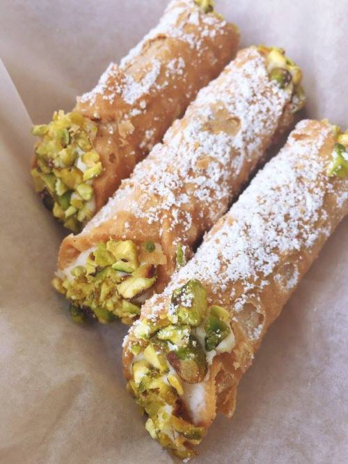 Three crispy cannoli filled with cream and topped with crushed pistachios and powdered sugar, resting on parchment paper.