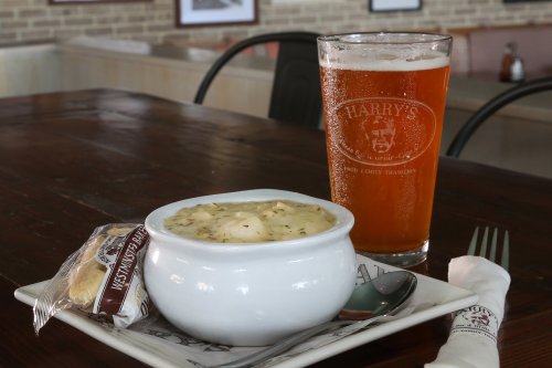 A bowl of soup with a biscuit and a glass of amber beer on a wooden table in a casual dining setting.