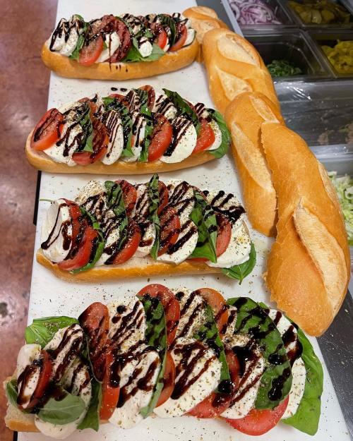 Four fresh sandwiches with mozzarella, tomatoes, basil, and balsamic drizzle, alongside sliced baguettes.