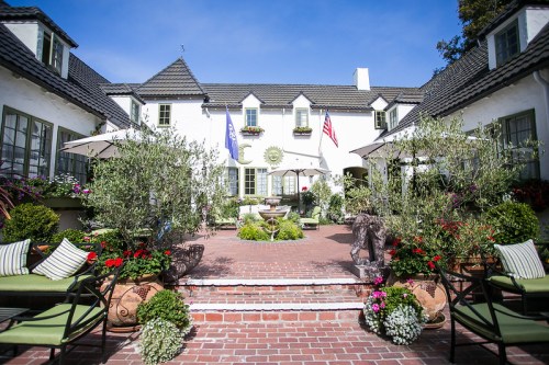 A charming courtyard with potted plants, flags, and seating, surrounded by a white building under a clear blue sky.