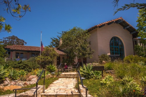 A charming library building surrounded by lush greenery and a stone pathway under a clear blue sky.