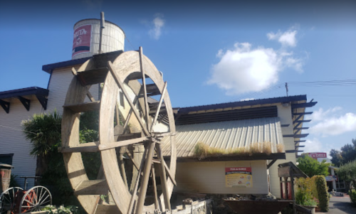 A large wooden water wheel in front of a building with a water tower and blue sky in the background.
