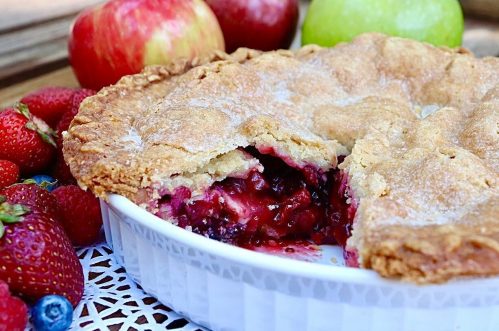 A freshly baked pie with a golden crust, filled with mixed berries, surrounded by apples and strawberries.