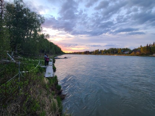 A serene river scene at sunset, with two people standing on a wooden dock surrounded by lush greenery.