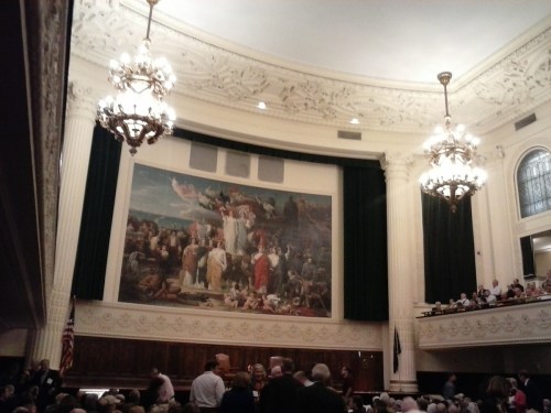 Interior of a grand hall with a large mural, ornate chandeliers, and an audience seated below.
