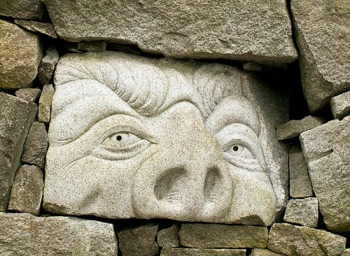 Carved stone face embedded in a wall of rough, gray stones, featuring exaggerated features and a prominent nose.