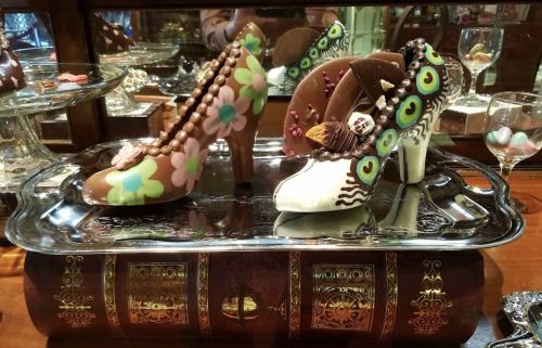 Two decorative chocolate high-heeled shoes on a silver tray, adorned with colorful designs and embellishments.