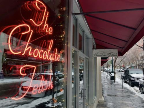 Neon sign for "The Chocolate Fetish" outside a storefront on a snowy street.